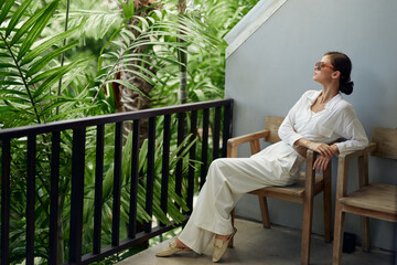Woman enjoying a morning on a tropical balcony, smiling and wearing trendy sunglasses She is...