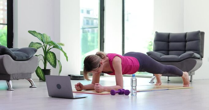 Fit woman does yoga plank pose following online lesson on mat maintaining body in light premise. Exercise to improve core of back and straight posture concept