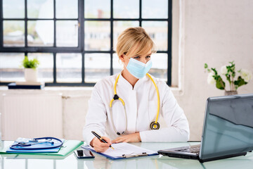 Female doctor with surgical mask sitting in doctor's room and using laptop for work