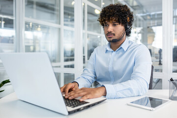 A young, professional man wearing a headset focuses intently on his laptop in a modern office environment, suggesting productivity and business communication.