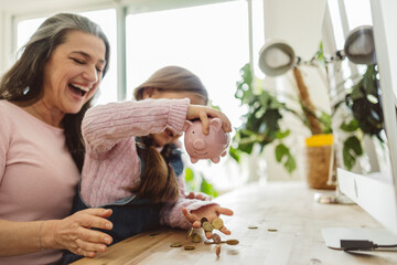 Granddaughter holding piggy bank over coins with grandmother at home