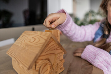 Girl inserting coin into wooden house shape bank at table at home