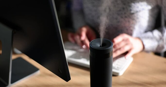 Female manager types on keyboard of computer sitting at table with aroma diffuser humidifier. Office worker enjoys scent of essential oils at workplace slow motion