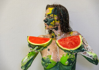 Portrait in profile of a sexy, yellow, green painted woman with large melons holding 2 fresh juicy red watermelon slices in her hands