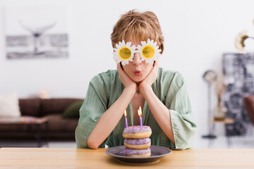 Happy woman in floral sunglasses blowing candles on doughnuts at home