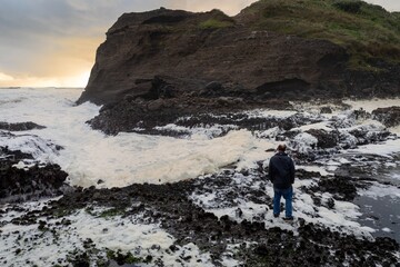 Man standing on rocks watching ocean waves and seafoam crashing over rocks at Piha, Auckland, New...