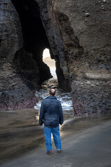 Man walking on a beach walking into a hole cave with waves coming through it. Piha, Auckland, New Zealand.