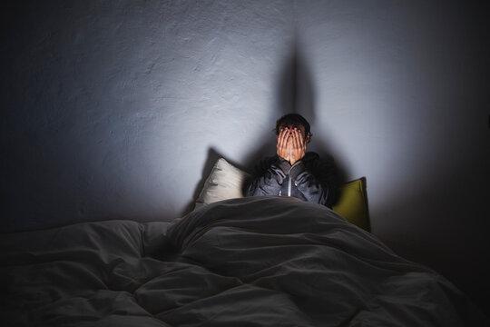 Depressed man covering face with hands sitting on bed in dark room