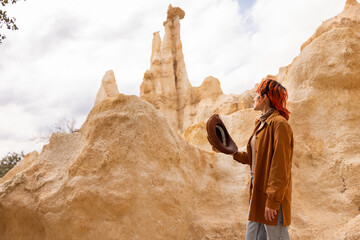A woman wearing a brown jacket and red hair is standing in front of a large rock formation. She is...