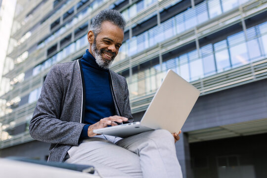 Smiling businessman working on laptop near building