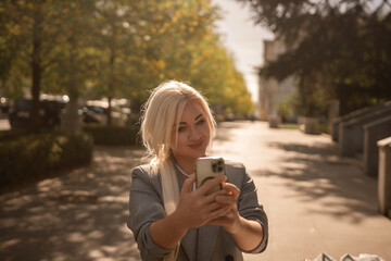 A woman is taking a picture of herself with her cell phone. She is wearing a gray jacket and scarf. The scene is set in a city with trees and cars in the background.