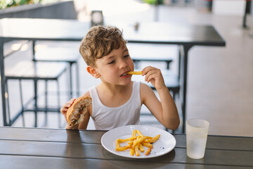 Little Boy Eating Sandwich and French Fries at Table. He appears focused on his meal, with a...