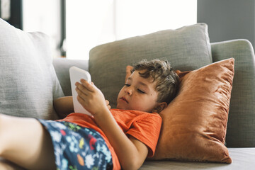 A boy with curly hair is reclining comfortably on a grey sofa with pillows, deeply immersed in the...