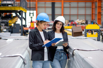 Two women with safety helmet and black suit standing in a factory, holding a clipboard and tablet computer, used for taking note, checking schedule, reviewing digital document, or communication.