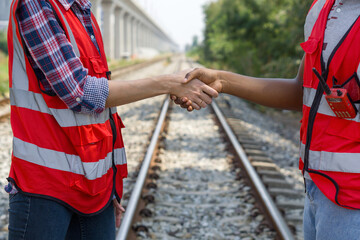 Two rail transportation engineer in reflective vest are shaking hands on some railroad tracks.