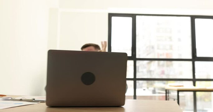 Positive male freelancer finishes work on laptop calling client via mobile phone. Office worker announces good news showing approval thumb up sign at table