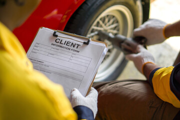 Closeup hand holding a clipboard featuring a form with client detail, ready to be filled out. A mechanic in uniform using a pneumatic impact wrench changing a tire in a background