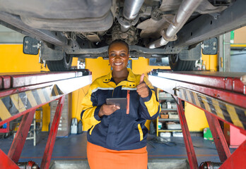 Young woman in work uniform, standing in a workshop with a car lifted on a hoist above her. The worker is smiling, holding a digital tablet in one hand and giving a thumbs-up with the other hand.