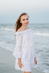 Portrait of a beautiful girl in a white dress. Female stand on the beach ocean and enjoy summer day on vacation. Happy woman go on seashore. Girl walks in water with big waves on sand sea in evening.