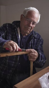 Older man preparing the wax frames for the hives in a workshop, pulling an iron with pliers, dressed in a blue checked shirt and standing in a workshop with natural light.
