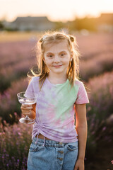 Young girl holding glass of water or lemonade in purple lavender field. Child enjoying summer time at orange sunset. Kid  among violet flowers with sunlight on spring day. Provence.