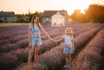 Mom with child girl on lavender field background. Mother holds hands little daughter in flowers lavender enjoying scent on summer day at sunset. Mom and baby embrace in meadow. Happy family outdoors.