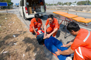Three paramedic or emergency medical technician (EMT) in orange uniform helping neck and head accident victim lying on stretcher long spinal board. Urgent assistance during road accident.
