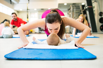 Mother smiling at baby while performing push-ups, kissing infant. Group exercise class, fitness or...