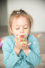 A sweet girl holds confetti in her hands and blows it away. She wears a bright smile as she enjoys the playful moment, creating an atmosphere of joy and excitement. The colorful confetti flies through