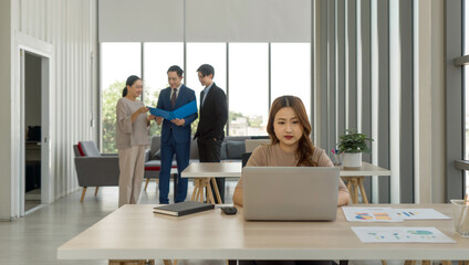 Young asian woman working with laptop computer on wooden table. In the background, three other individuals are engaged in a conversation, sharing a document. Atmosphere in office with large window.