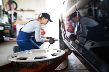 Female auto mechanic changing tieres in auto service. Beautiful woman kneeling by wheel in a garage, wearing blue coveralls.