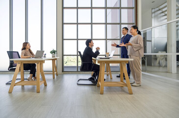 Four professional in a modern office with large window talk and work at wooden desk.