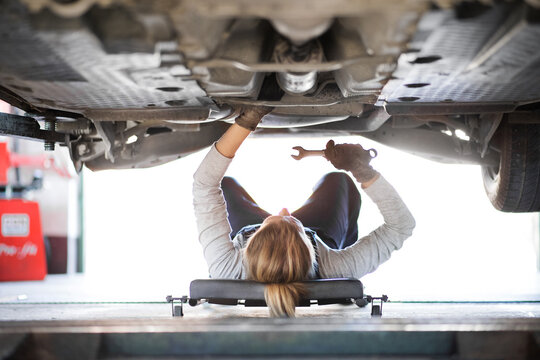 Female auto mechanic lying on mechanic creeper under car, inspecting and repairing vehicle. Beautiful woman working in a garage, wearing blue coveralls.
