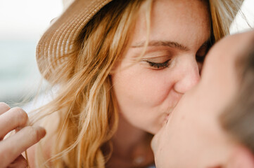 Closeup face. Male kisses female sitting on beach ocean and enjoys sunny summer day. Man embraces...