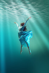 Under the sea surface. Young girl in elegant dress levitating underwater, extending hands toward sunlight filtering through water. Concept of surrealism, beauty, mystery and fantasy, freedom