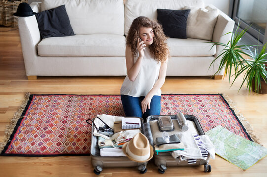 Beautiful woman planning summer vacation abroad, sitting on floor, packing suitcase. Solo traveler making phone call.