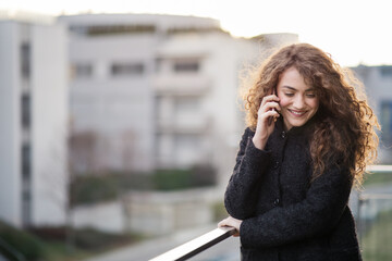 Beautiful woman with curly hair standing on terrace, making phone call outdoors during cold spring...