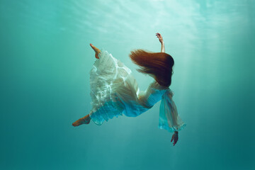 Young woman in flawless dress, with red hair captured in the tranquility of deep water, diving...