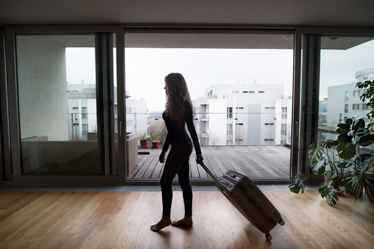 Woman is pulling wheeled travel suitcase behind her, leaving the apartment for summer vacation. Solo traveller going on trip.