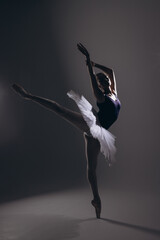 Young ballerina in elegance white tutu and pointe shoes performing Arabesque pose against dark...