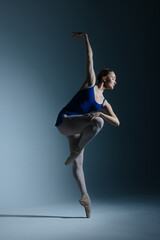Young ballerina in bodysuit and pointe shoes dancing against of blue background.