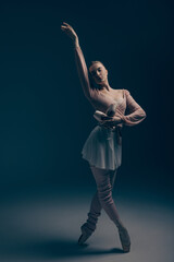 Young ballerina in dress and pointe shoes dancing against blue background.