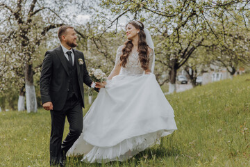 the groom and his bride are walking on the green grass in the spring garden. The bride is in a chic white dress, the groom is in a black suit