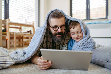 Little boy watching cartoon movie on tablet with father, lying under blanket on floor in kids room. Dad explaining technology to son, digital literacy for kids.