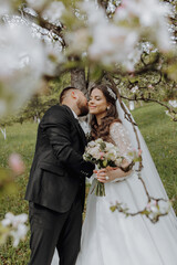 The groom kisses his bride in a white dress against the background of blooming trees in a green garden in spring