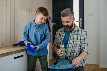 Father helping son get ready for kindergarten, preschool. Putting lunchbox with food, snacks in...
