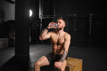 Man resting after exercise, drinking water from bottle, sitting, wearing short with bare chest....