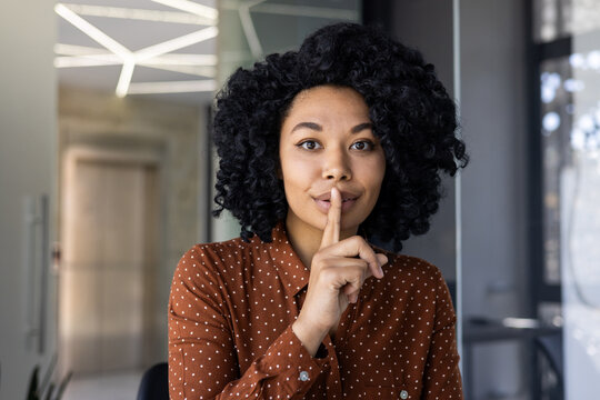 African American woman with curly hair making a shushing gesture, signaling silence in a contemporary office environment. Her playful expression conveys secrecy or surprise.
