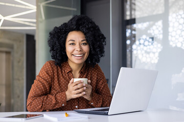A cheerful young African American businesswoman enjoys a coffee break while working on her laptop...