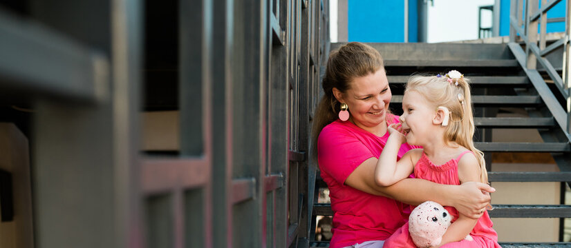 Blonde little girl with cochlear implant playing with her mother outdoor. Hear impairment deaf and health concept. Disability and inclusion. Copy space